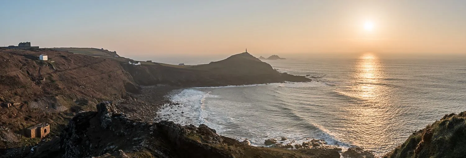 Panoramic view of a rocky coastline with a headland, scattered buildings, and the sun setting over the sea.