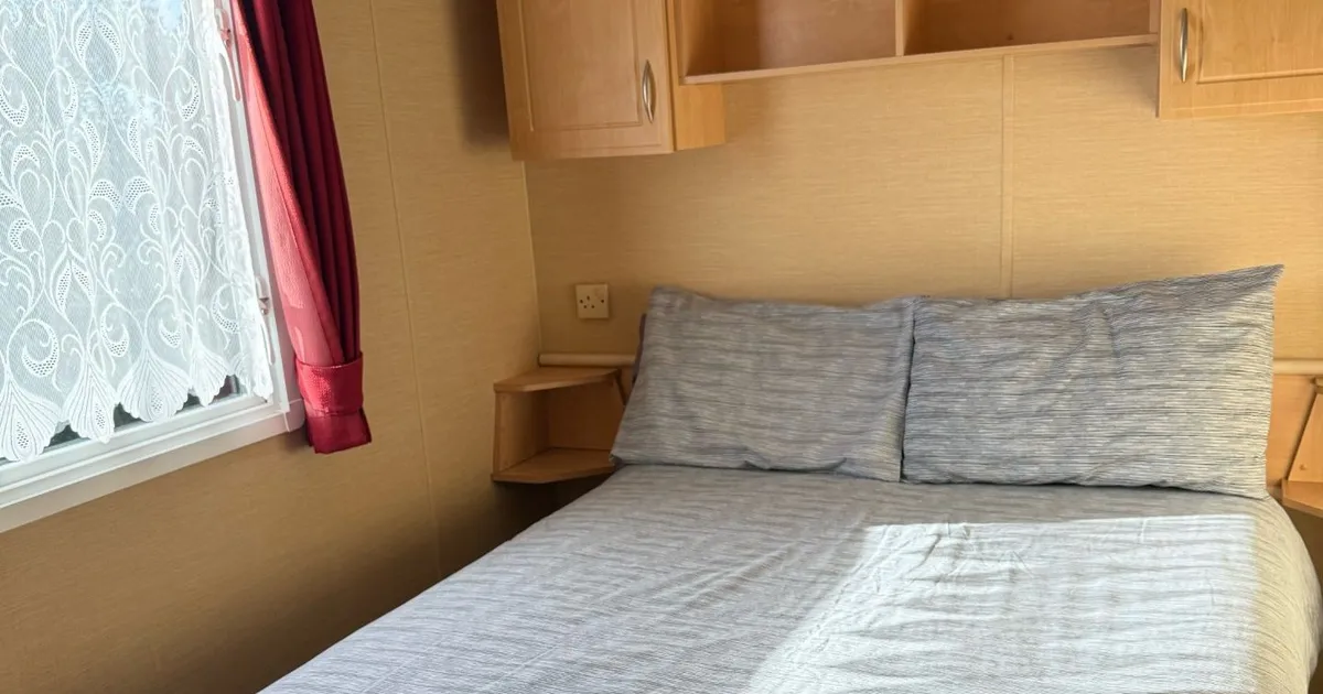 A neatly made bed in a small bedroom with wooden cabinets, shelves, and a window with red curtains.