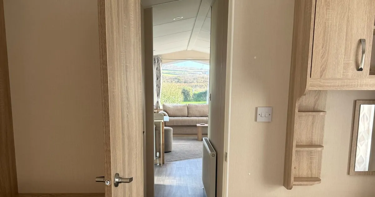 Interior view of a cozy mobile home or caravan bedroom looking through an open door into a living area with wooden furniture and a view outside.