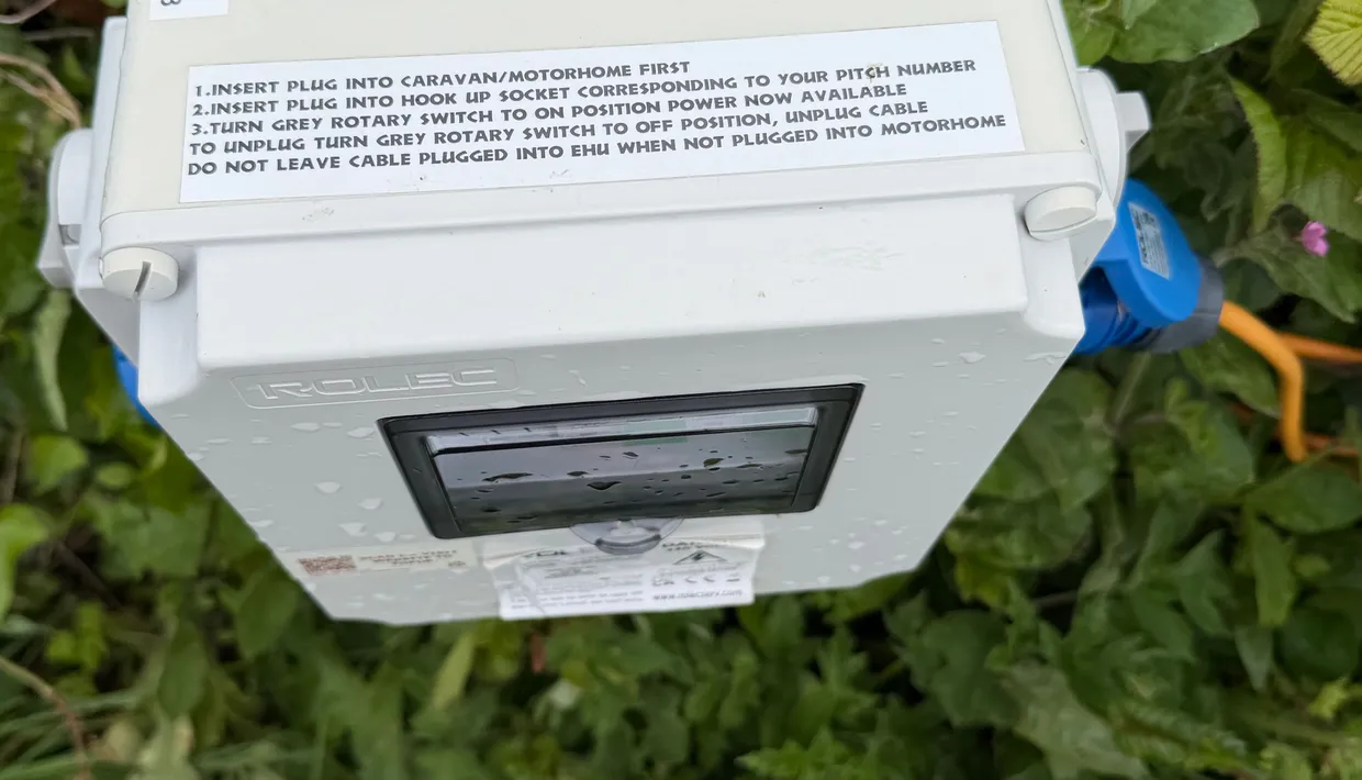 Outdoor caravan electric hook-up box with top-up instructions among green leaves