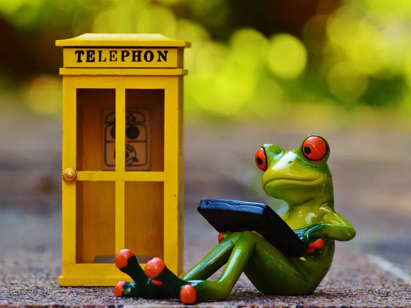 Green toy frog with red eyes holding a smartphone beside a yellow telephone box