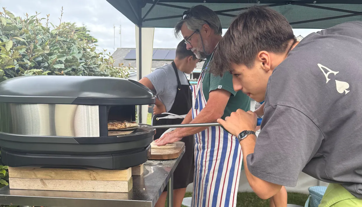 Group of people making pizza outdoors under a canopy tent with a portable pizza oven.