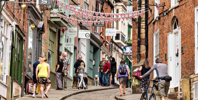 People stroll and shop along a steep cobbled street lined with historic brick buildings, Union Jack bunting, and quaint shopfronts.