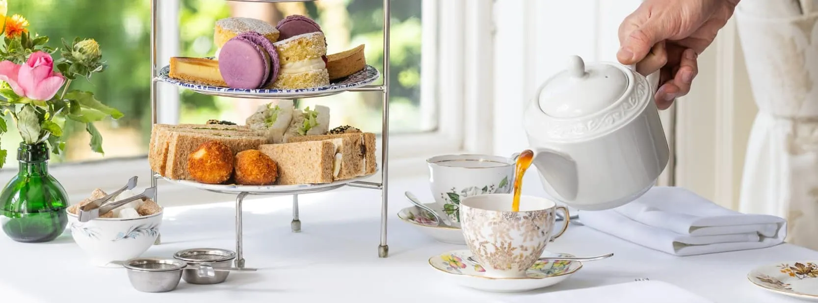 A person pours tea from a white teapot into a floral cup at a beautifully arranged afternoon tea table with pastries, sandwiches, and fresh flowers.