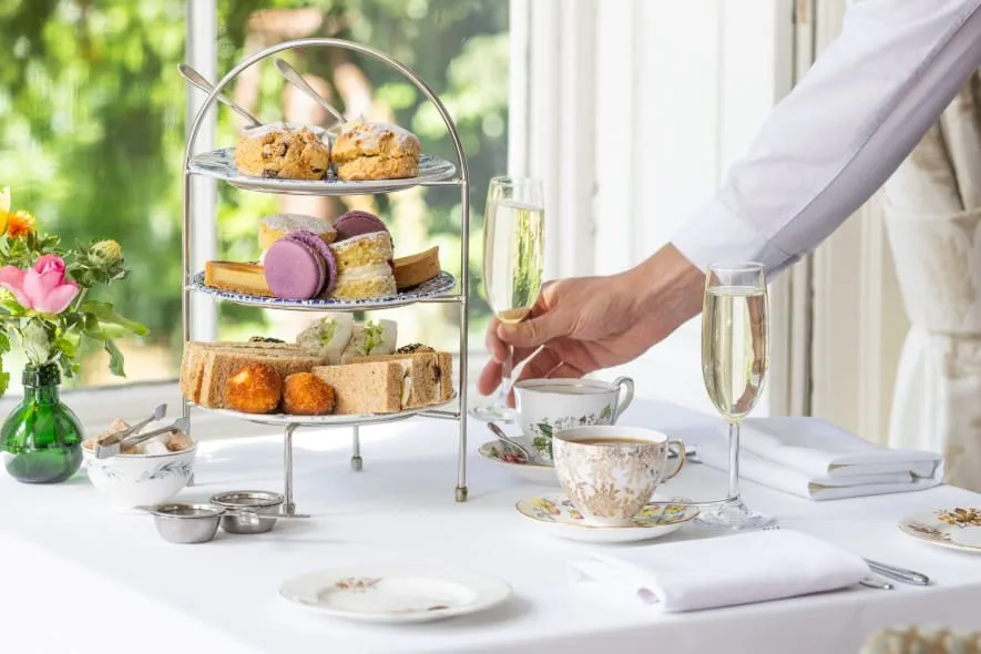 A server in a white shirt places a champagne flute on a table set for afternoon tea with tiered treats, fine china, and fresh flowers near a sunlit window.