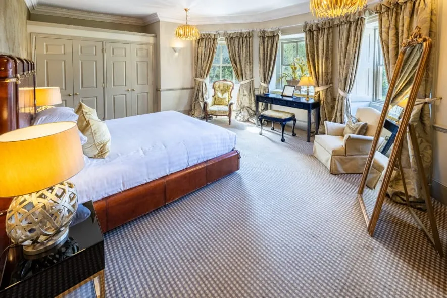 Bright, traditional-style bedroom with a large bed, plush carpet, gold-toned accents, and a wall of windows dressed in patterned curtains.