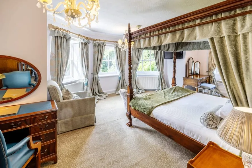 Corner view of a stately bedroom with a canopy bed, chandelier, writing desk, and tall windows draped in floral curtains.