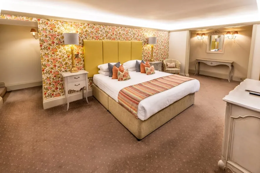 Country-house bedroom with super king bed, yellow headboard, floral wallpaper, bedside lamps and dressing table.
