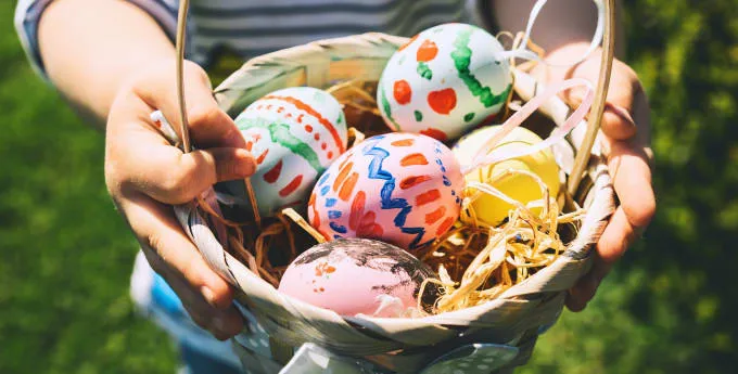 An image of a basket being held in a pair hands, which is full of colourfully painted easter eggs with straw underneath.