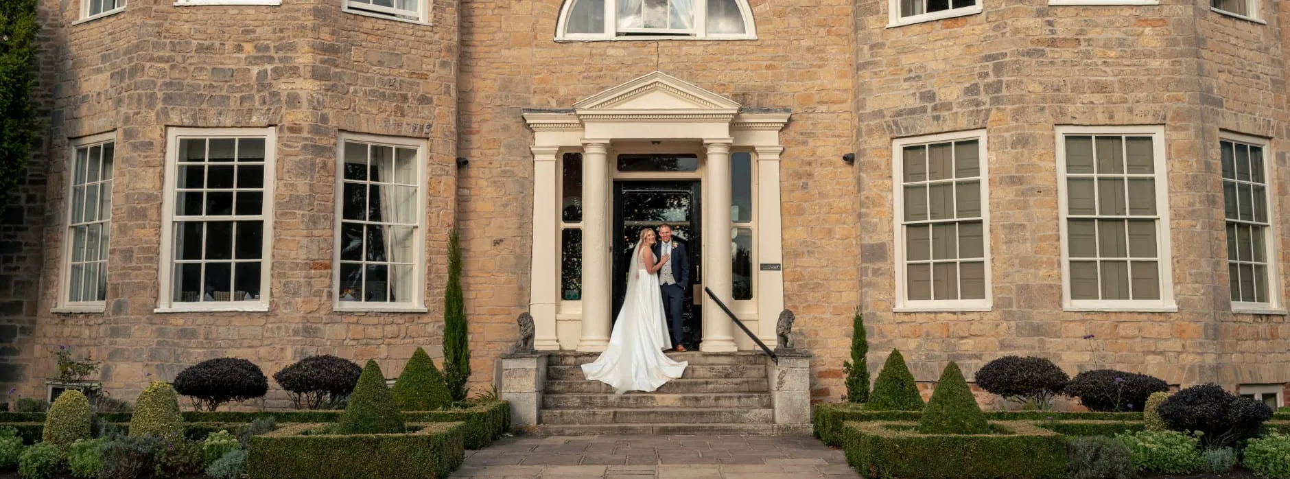 Bride and groom stand in front of a grand stone manor house entrance, smiling together on the steps beneath elegant columns.