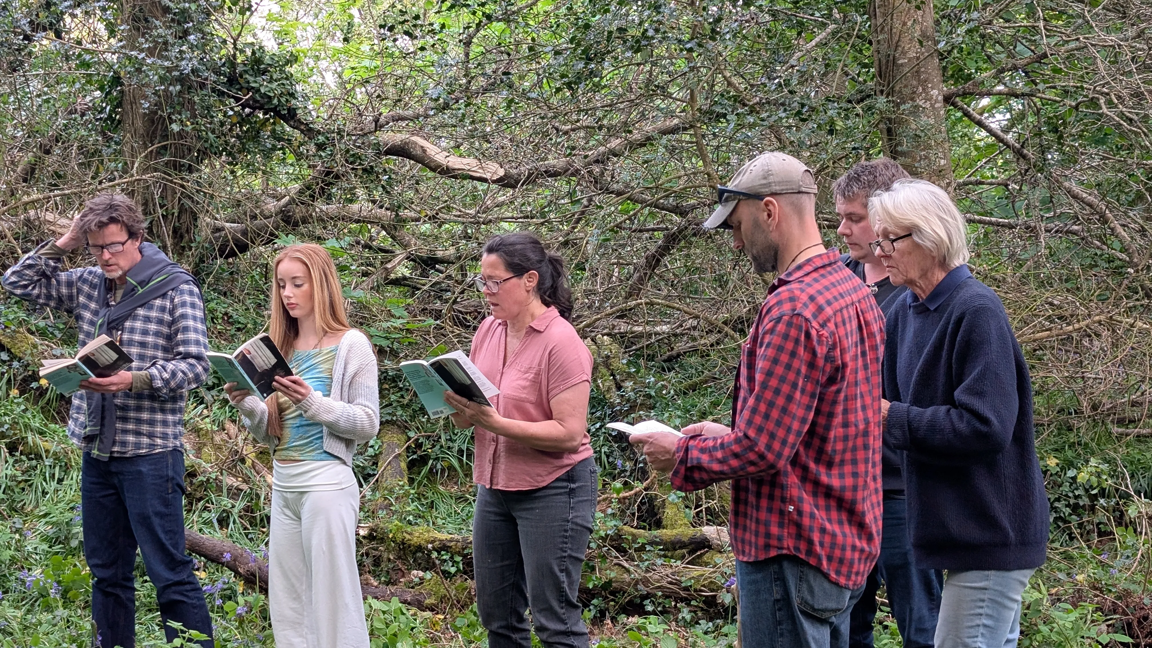 Cast members rehearse The Crucible outdoors in a wooded area, standing in a line and reading from scripts.