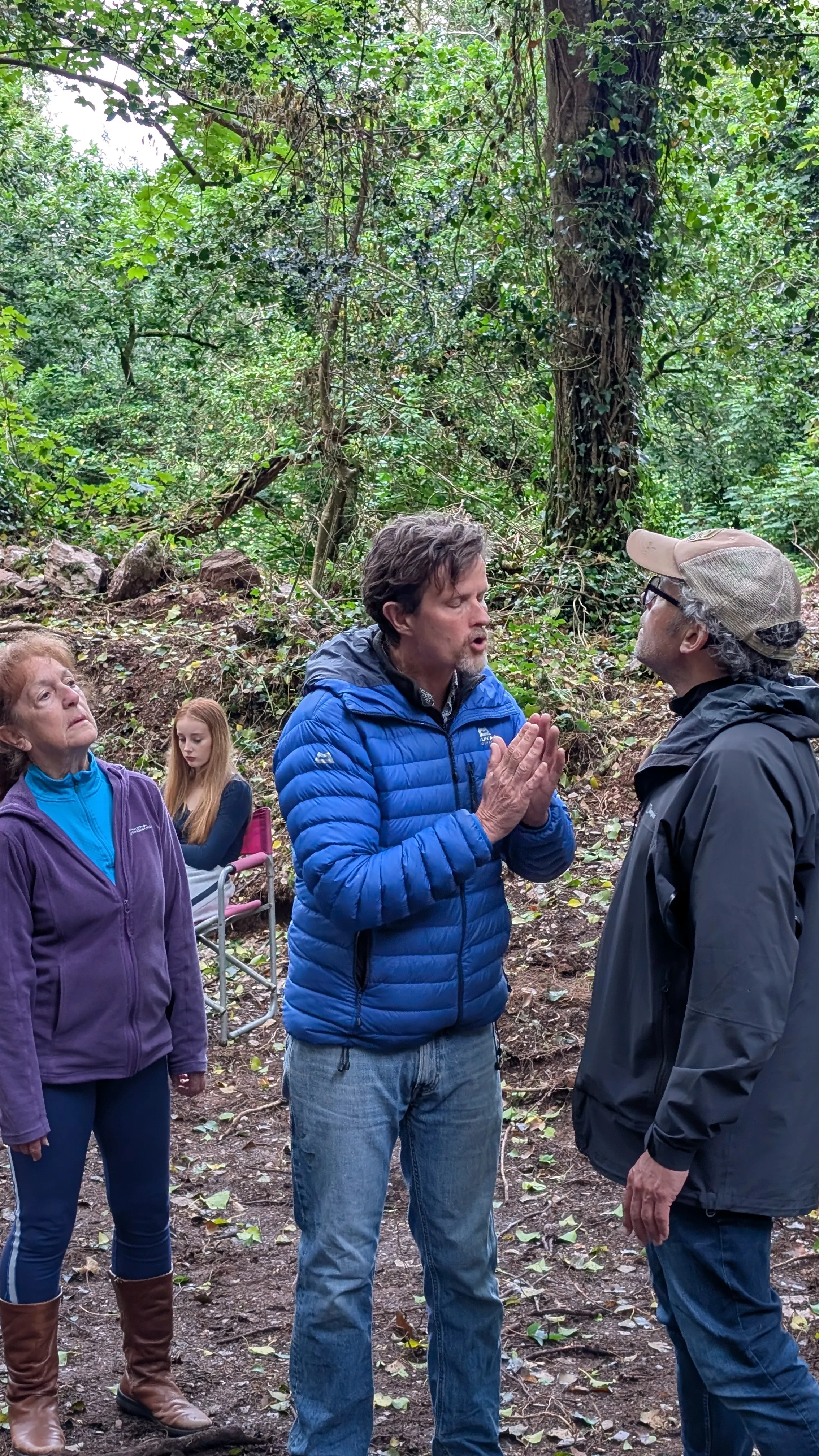 Three performers and a crew member stand in a wooded clearing during an outdoor rehearsal of The Crucible, with one seated cast member in the background.