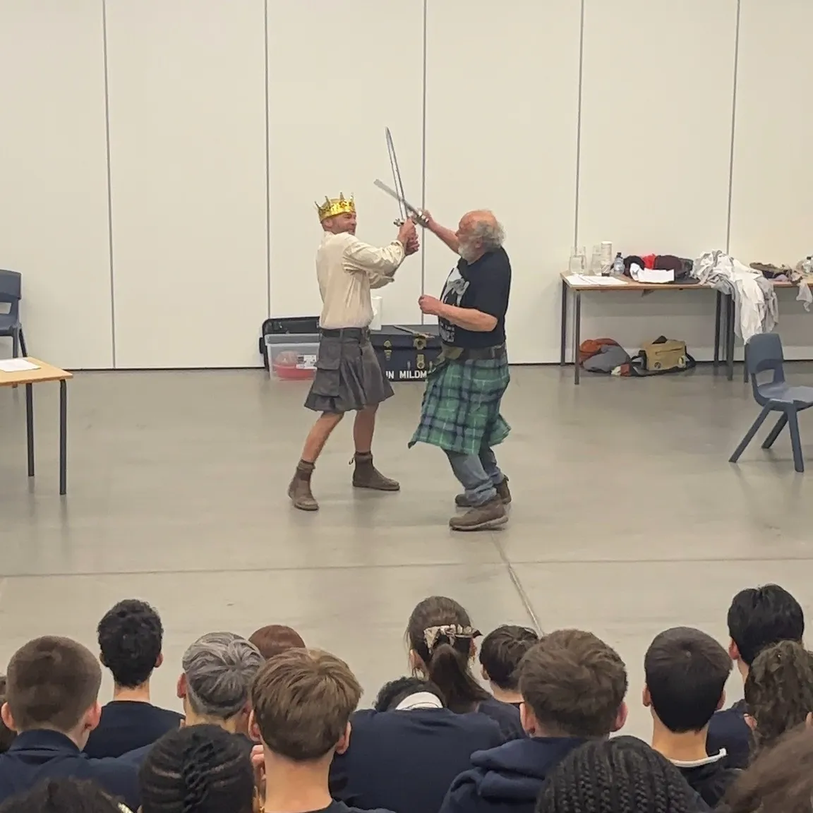 Two men in kilts perform a staged sword fight in front of a seated audience in a hall, with one wearing a gold crown.