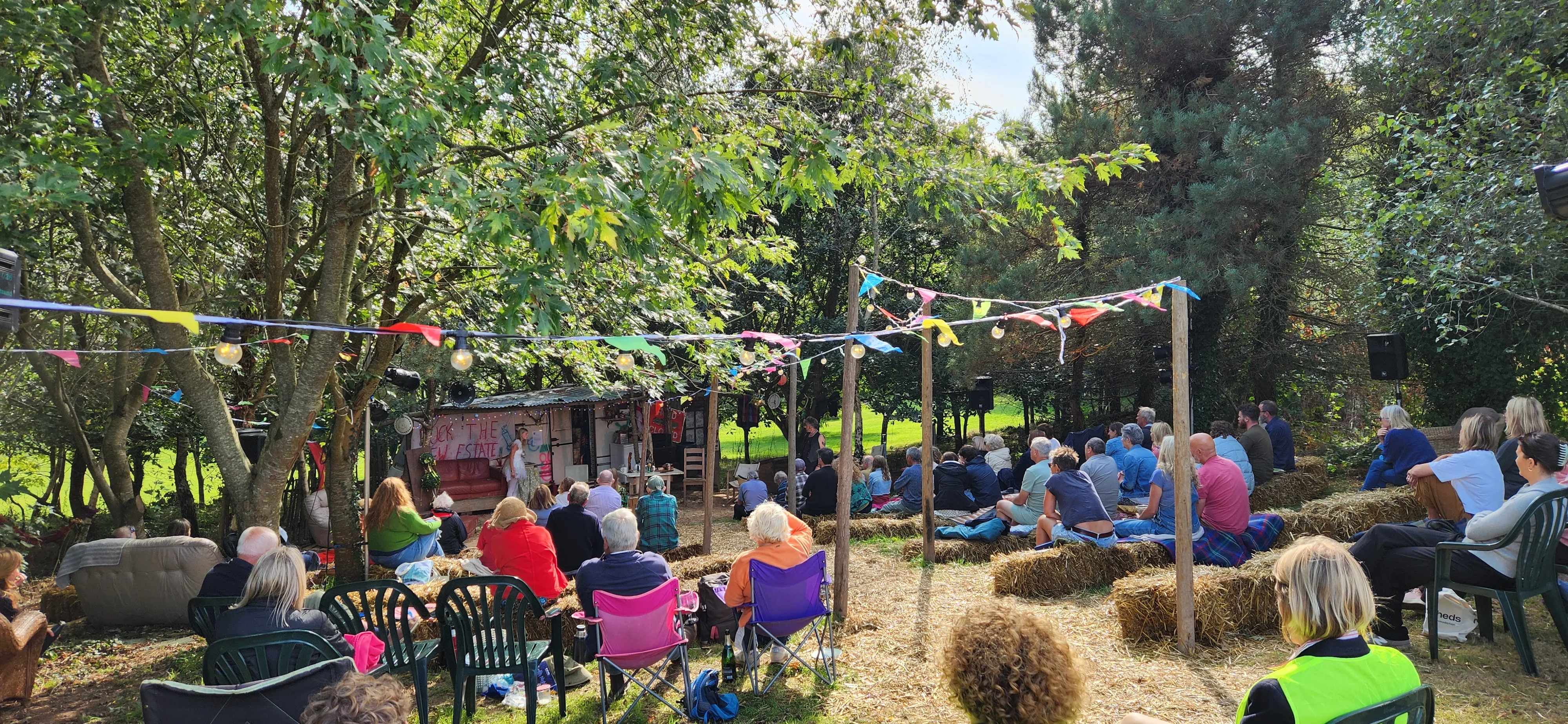 People sitting on hay bales and chairs at an outdoor event under colorful bunting in a wooded area.