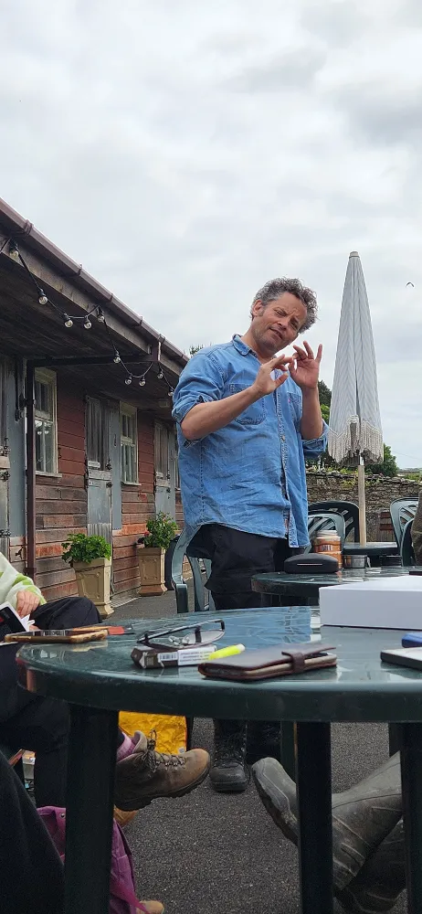 Man in a blue shirt gesturing with his hands while standing outside near a table, with a rustic building and an umbrella in the background.