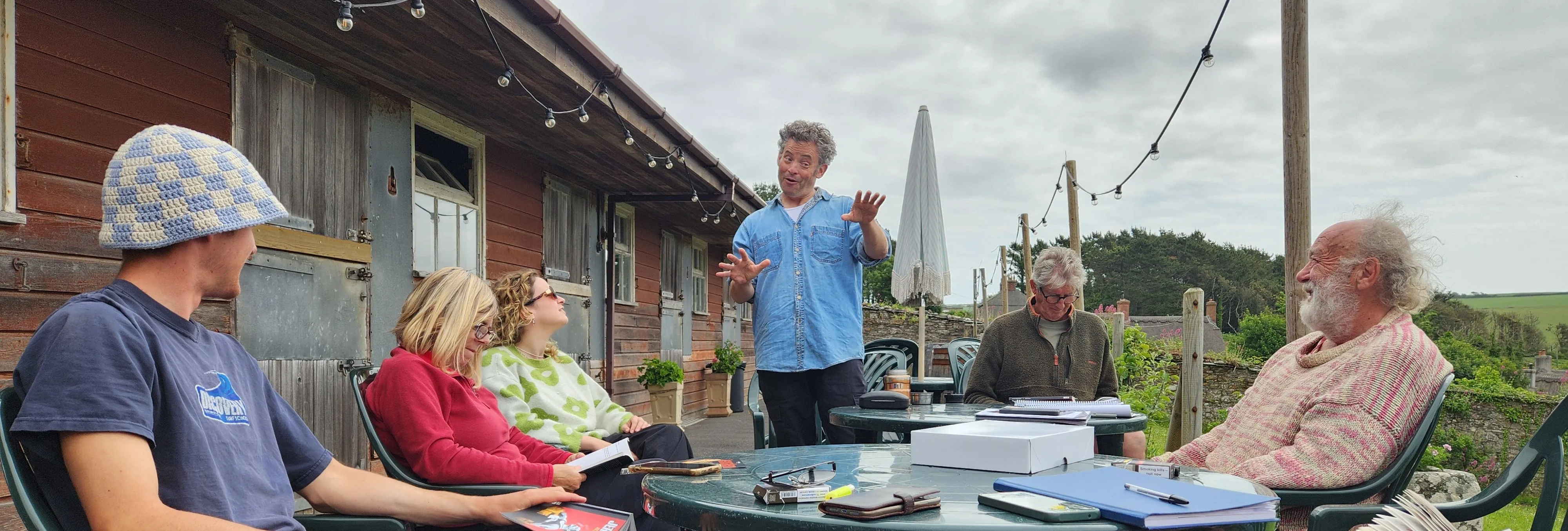 A group of people sitting outside around a table, engaged in a lively discussion or meeting, with one person standing and gesturing as they speak.