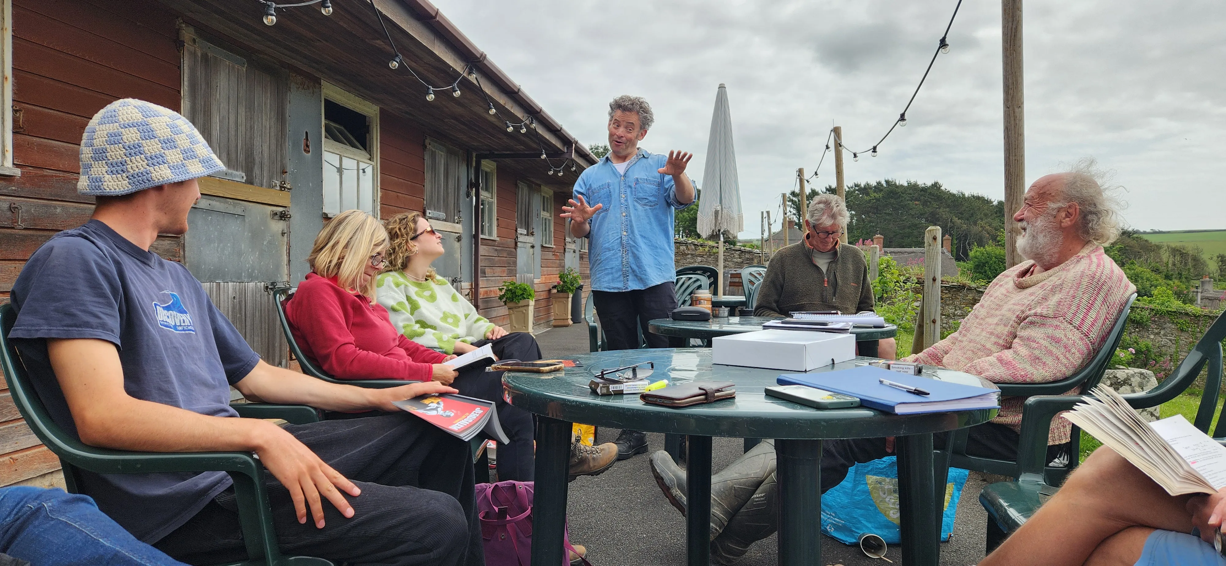 A group of people sitting outside around a table, engaged in a lively discussion or meeting, with one person standing and gesturing as they speak.