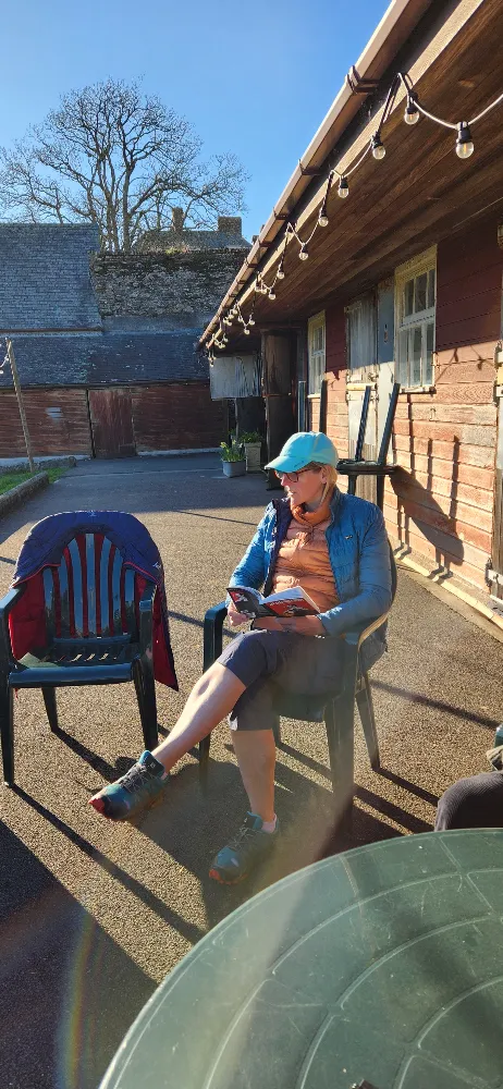 Person sitting outside in the sun, wearing a blue cap and jacket, reading a book beside a table and another empty chair with a jacket draped over it.
