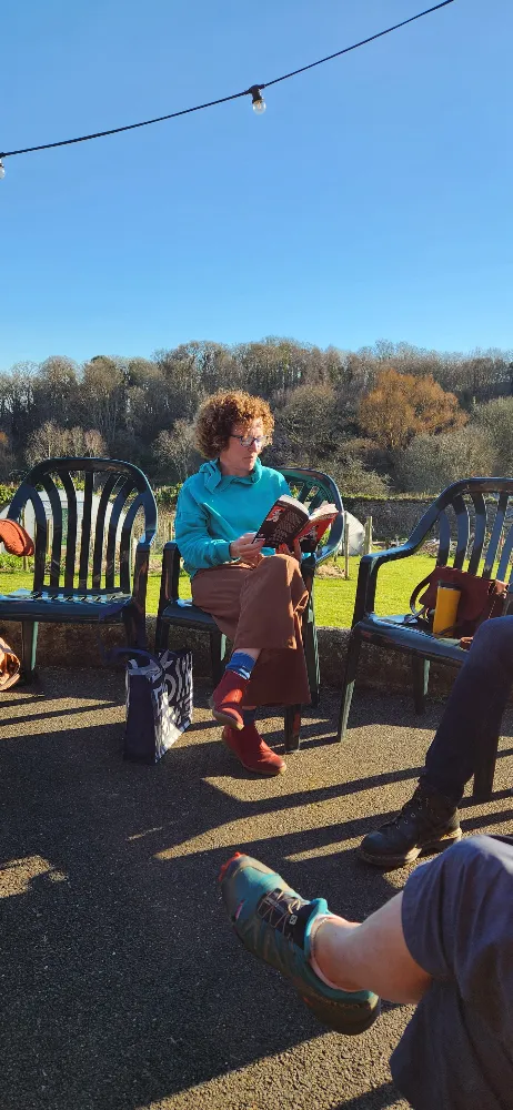 Person sitting on a plastic chair outdoors, reading a book, surrounded by others on chairs, with a background of trees and blue sky.