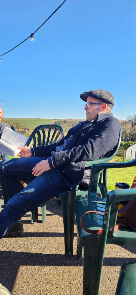 Man in a flat cap and glasses sitting on a green plastic chair outdoors, reading papers, with a blue sky and countryside in the background.