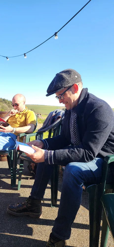Two men sitting on outdoor chairs reading in the sunshine, string lights overhead and a clear blue sky