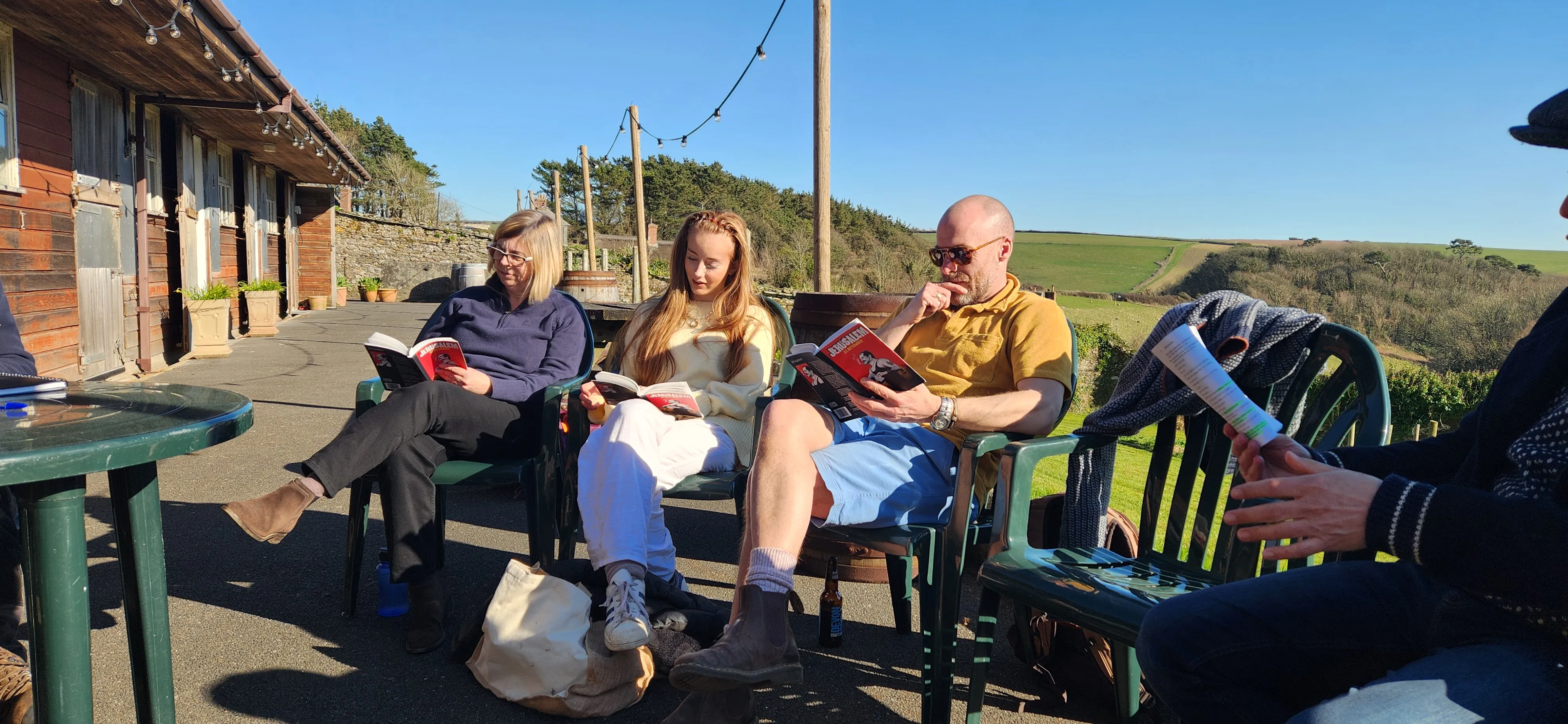 People sitting outdoors on plastic chairs reading books on a sunny day near a rustic building and fields.