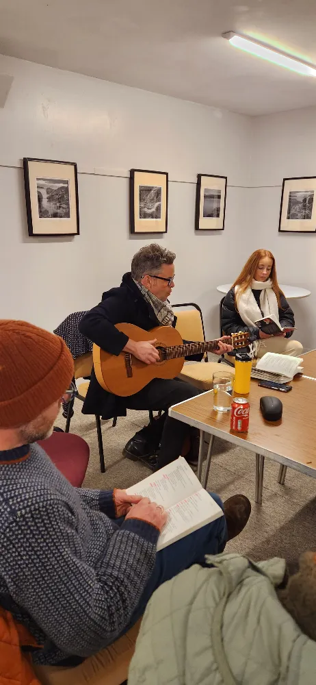 A man playing guitar in a small room with people reading books around a table.