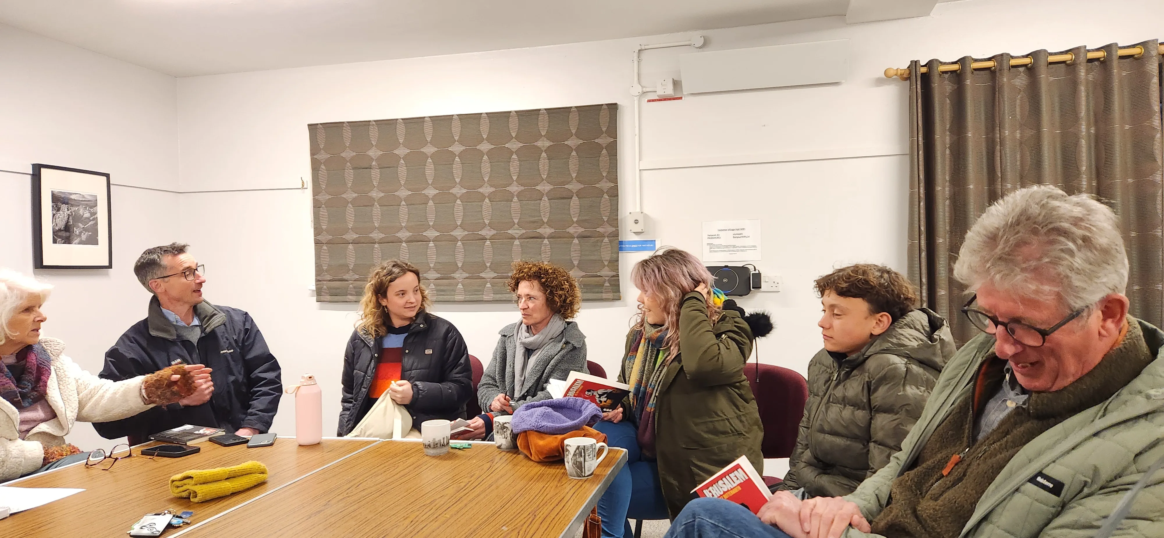 A group of people sitting around a table in a brightly lit room, engaged in discussion. Some are holding books and mugs, suggesting a book club meeting.