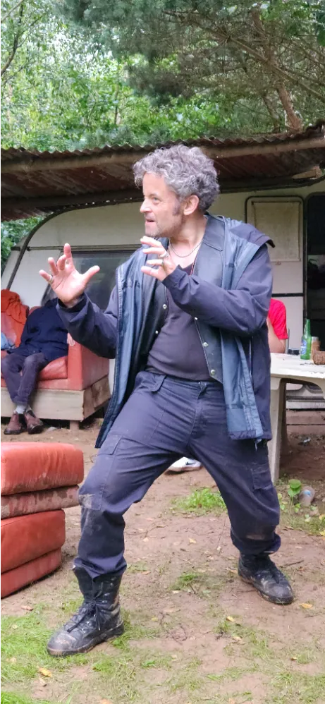Man with curly gray hair gesturing expressively outdoors in front of a caravan