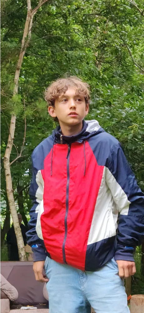 Teenager wearing a red, navy, and white jacket standing outdoors in a wooded area.