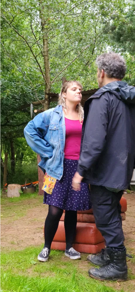 Two people standing and facing each other outdoors, one with hands on hips wearing a denim jacket.