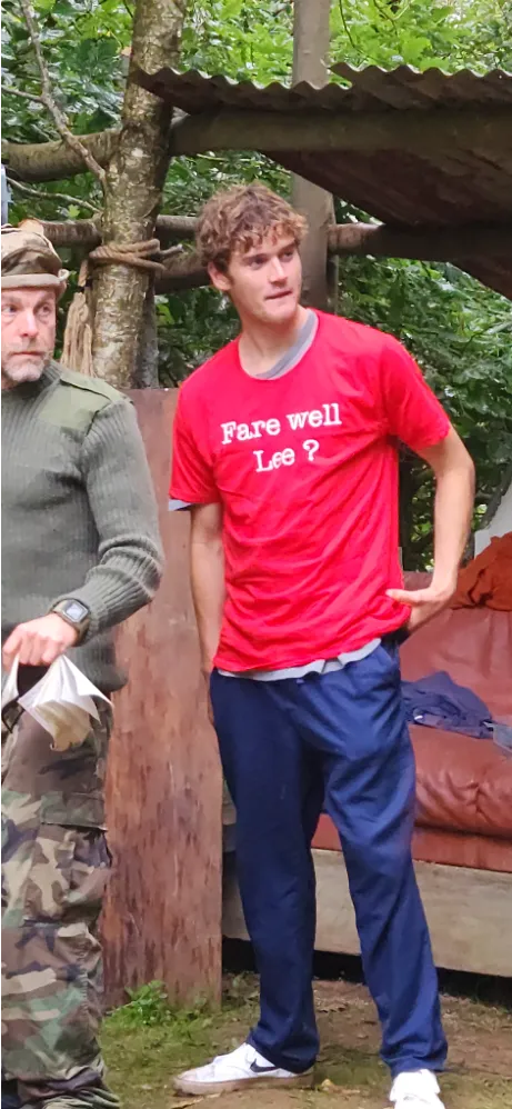 Young man in red t-shirt with 'Fare well Lee?' and blue pants standing outdoors near another person in camouflage.