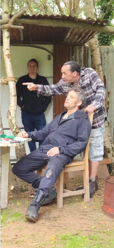 Three men outside near a rustic shelter, one sitting on a chair with eyes closed, another standing beside him pointing, and a third man in the background.