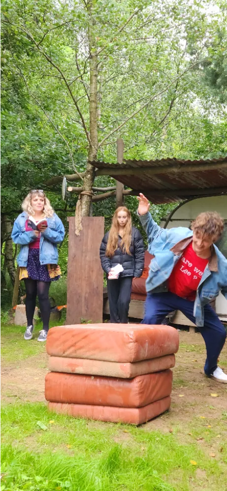 Three people outdoors near trees; one is reading, one is standing, and one is leaning forward near a stack of cushions.