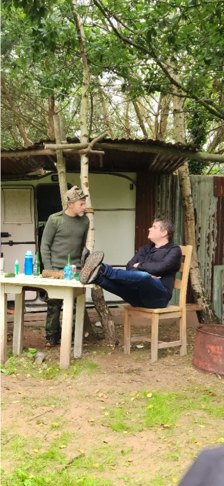 Two men sitting and talking at a table outdoors, surrounded by trees and rustic structures.