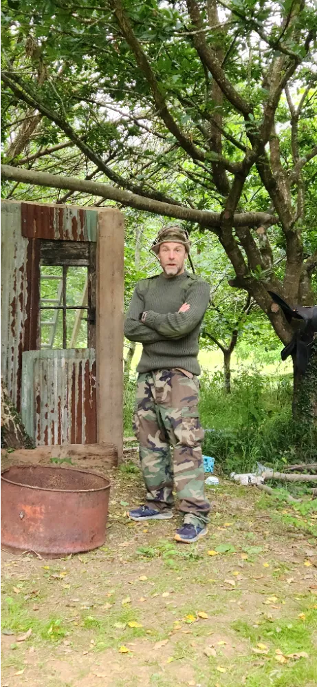 Man in camouflage pants and green sweater standing outdoors near a rustic structure and trees