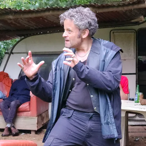 Man gesturing dramatically outdoors near a table and an old couch.
