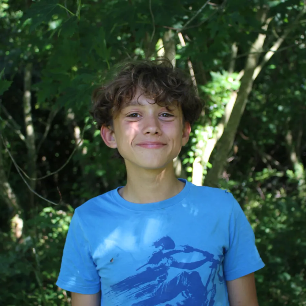 Smiling boy with curly hair wearing a blue t-shirt standing outdoors in a wooded area.