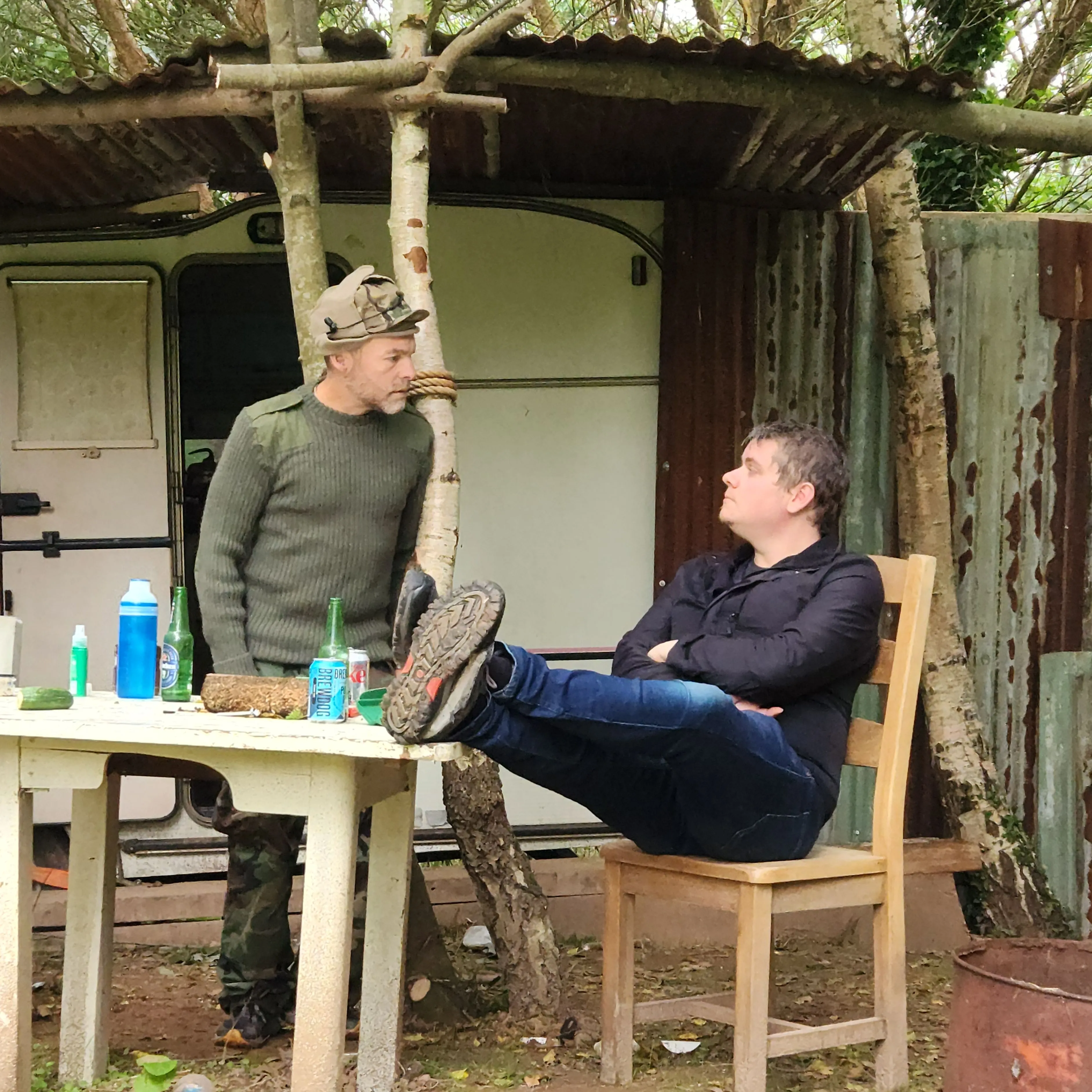Two men talking at an outdoor table in a rustic, wooded setting with a shed and a caravan in the background.