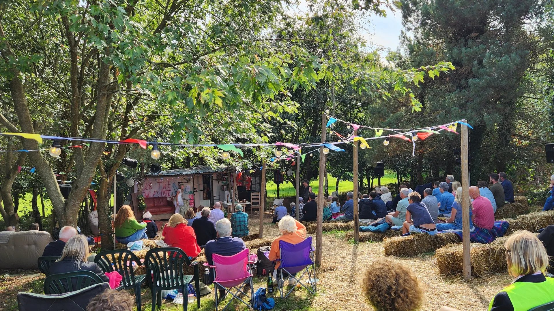People sitting on hay bales and chairs at an outdoor event under colorful bunting in a wooded area.