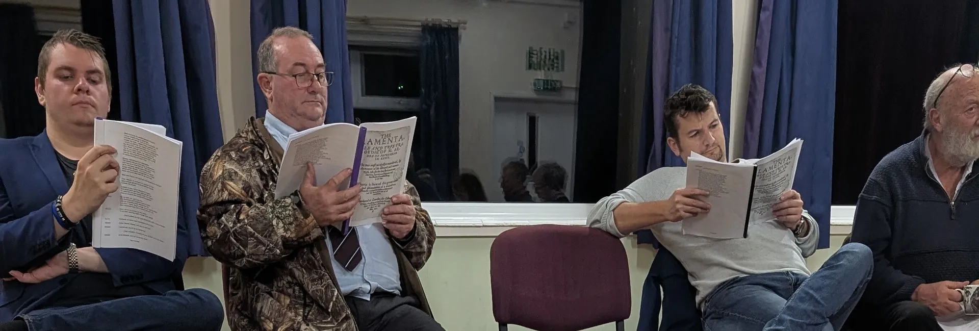 A group of people seated in a semicircle, each holding a script and reading, with an open script in the foreground out of focus.