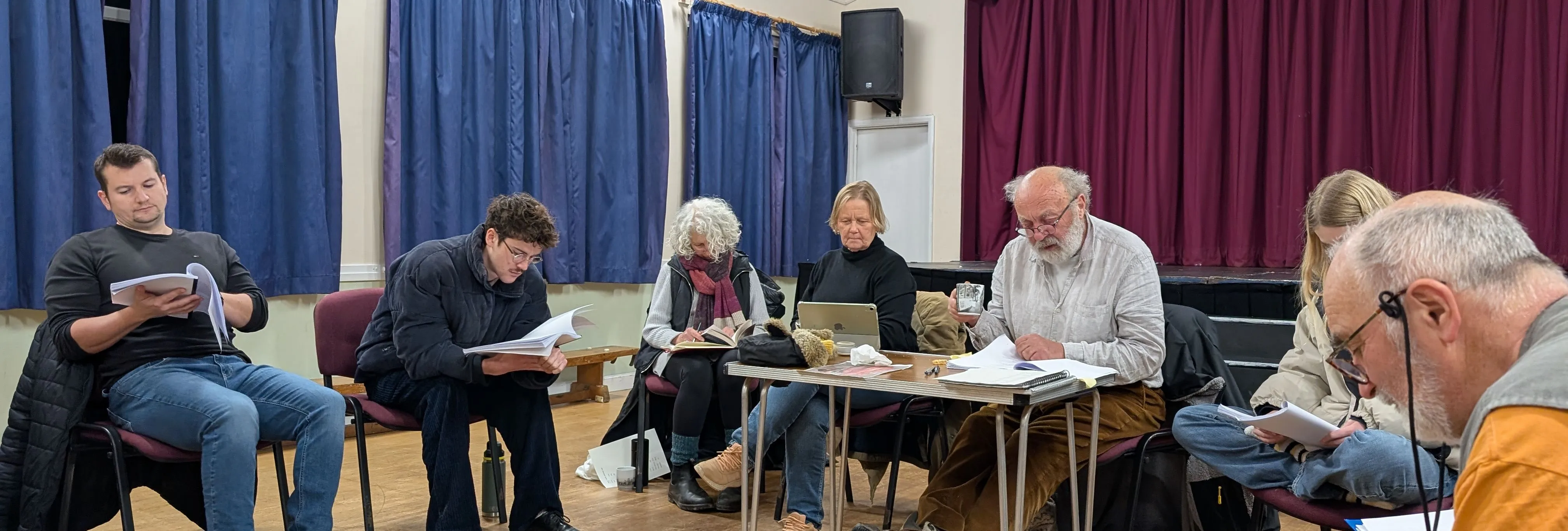 A group of people sitting in a circle reading scripts in a room with blue curtains and a small stage.