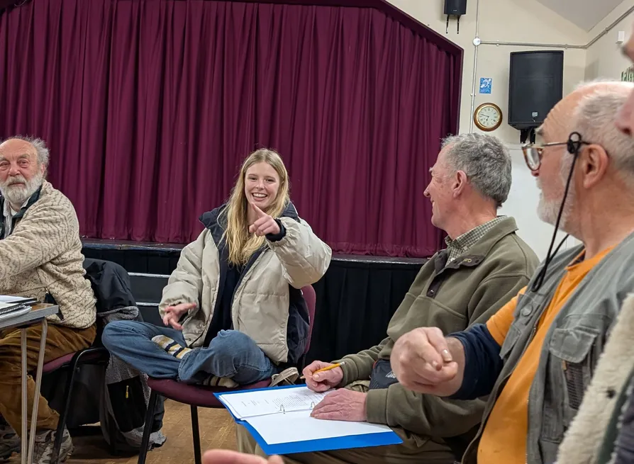 Group of people sitting in a room with a stage and maroon curtain in the background. One young woman is smiling and pointing, others are engaged in conversation.