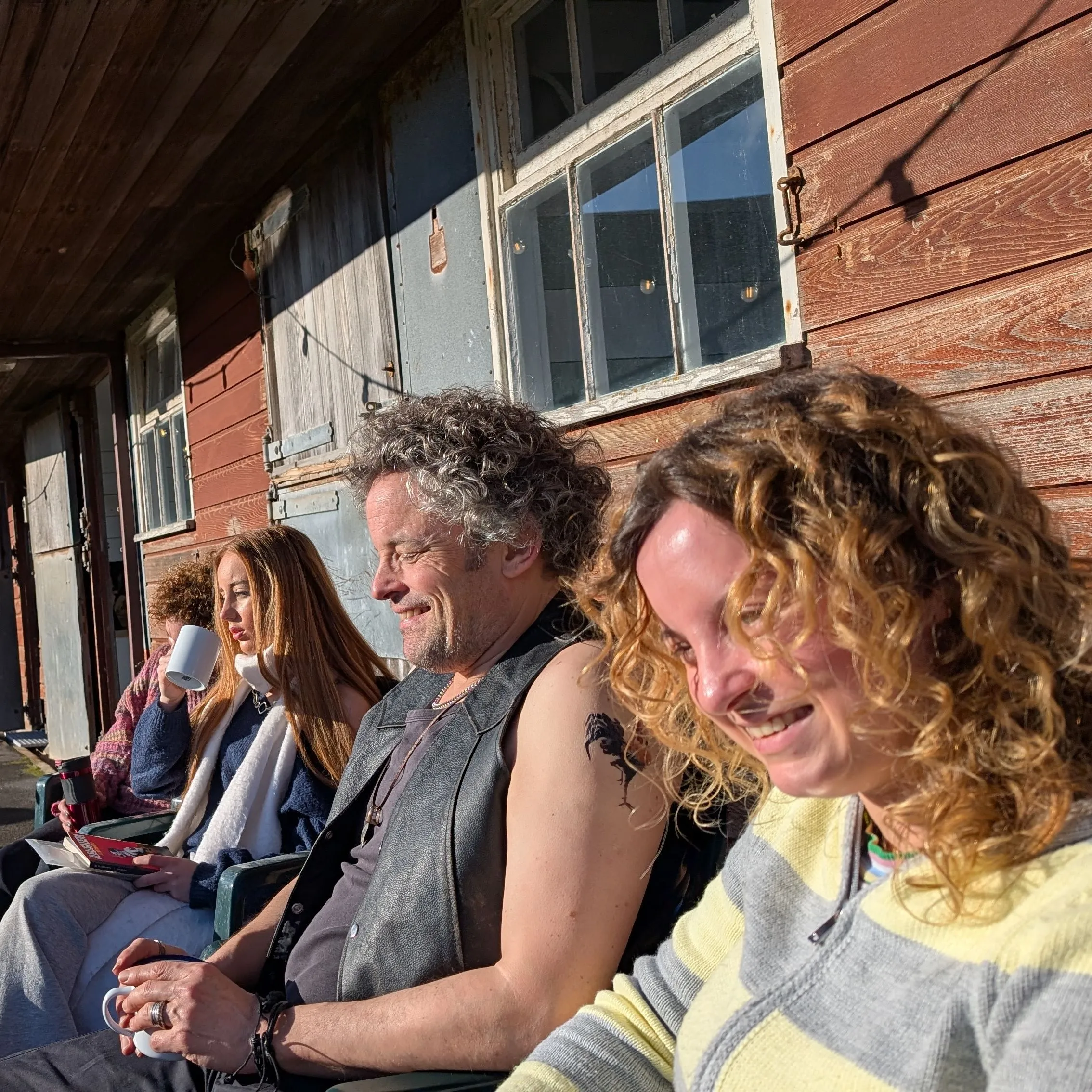 Group of people sitting outside on chairs next to a rustic wooden building, enjoying sunny weather.