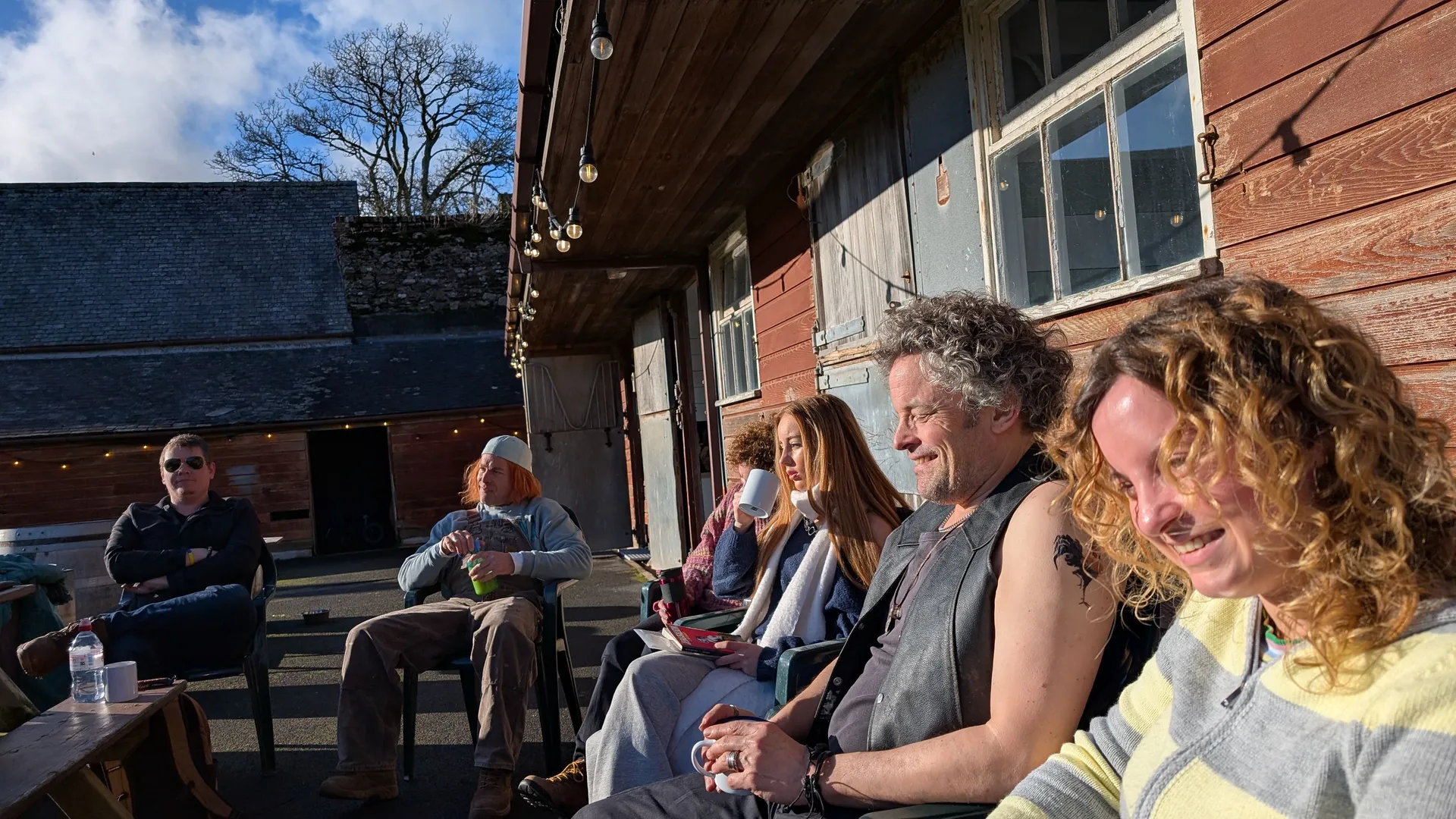 Group of people sitting outside on chairs next to a rustic wooden building, enjoying sunny weather.