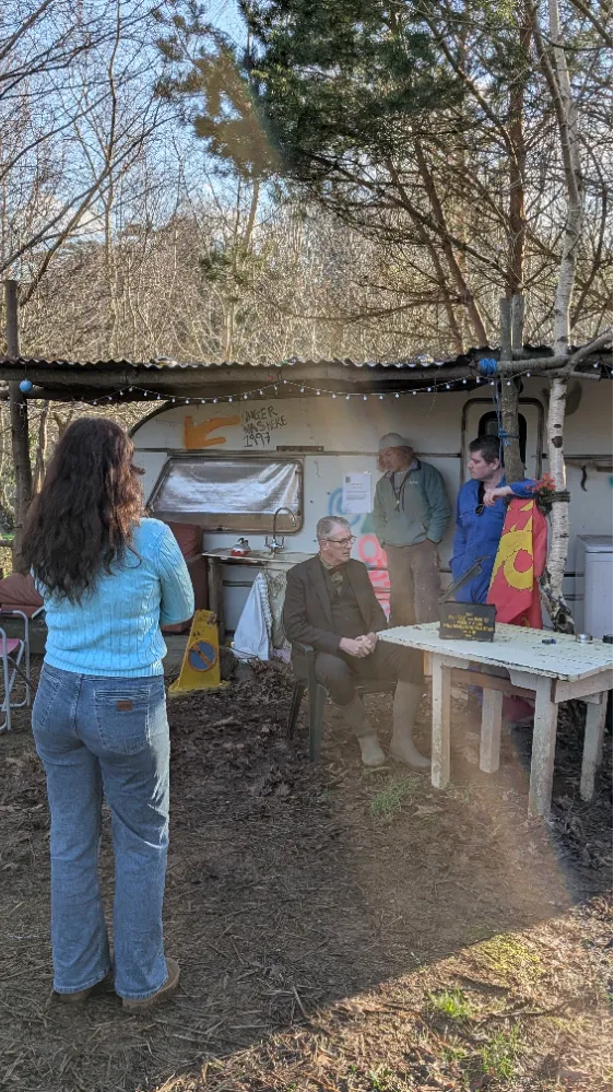 Group of people outdoors at a rustic campsite with a table, chairs, and a caravan in the background.