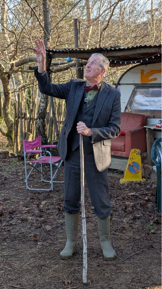 Elderly man in a grey suit and bow tie, wearing rubber boots, holding a stick and gesturing upwards outdoors near a camper.