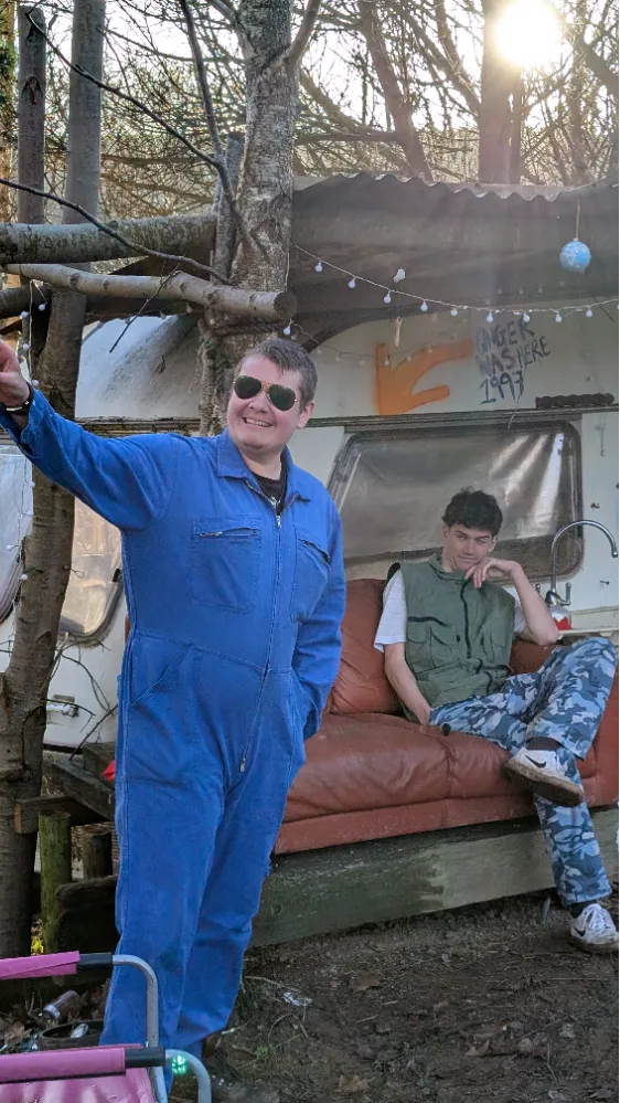 Two people outdoors, one in a blue jumpsuit and sunglasses smiling and pointing, another sitting on a brown couch in front of a caravan.