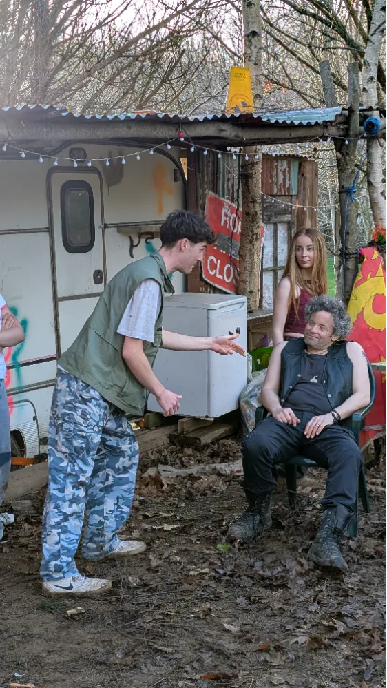 A group of people outside a rustic caravan, with one man sitting and others standing, surrounded by trees and outdoor decor.