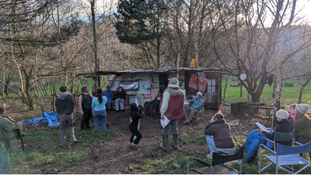 A group of people gathered outdoors near a makeshift shelter or caravan in a wooded area.
